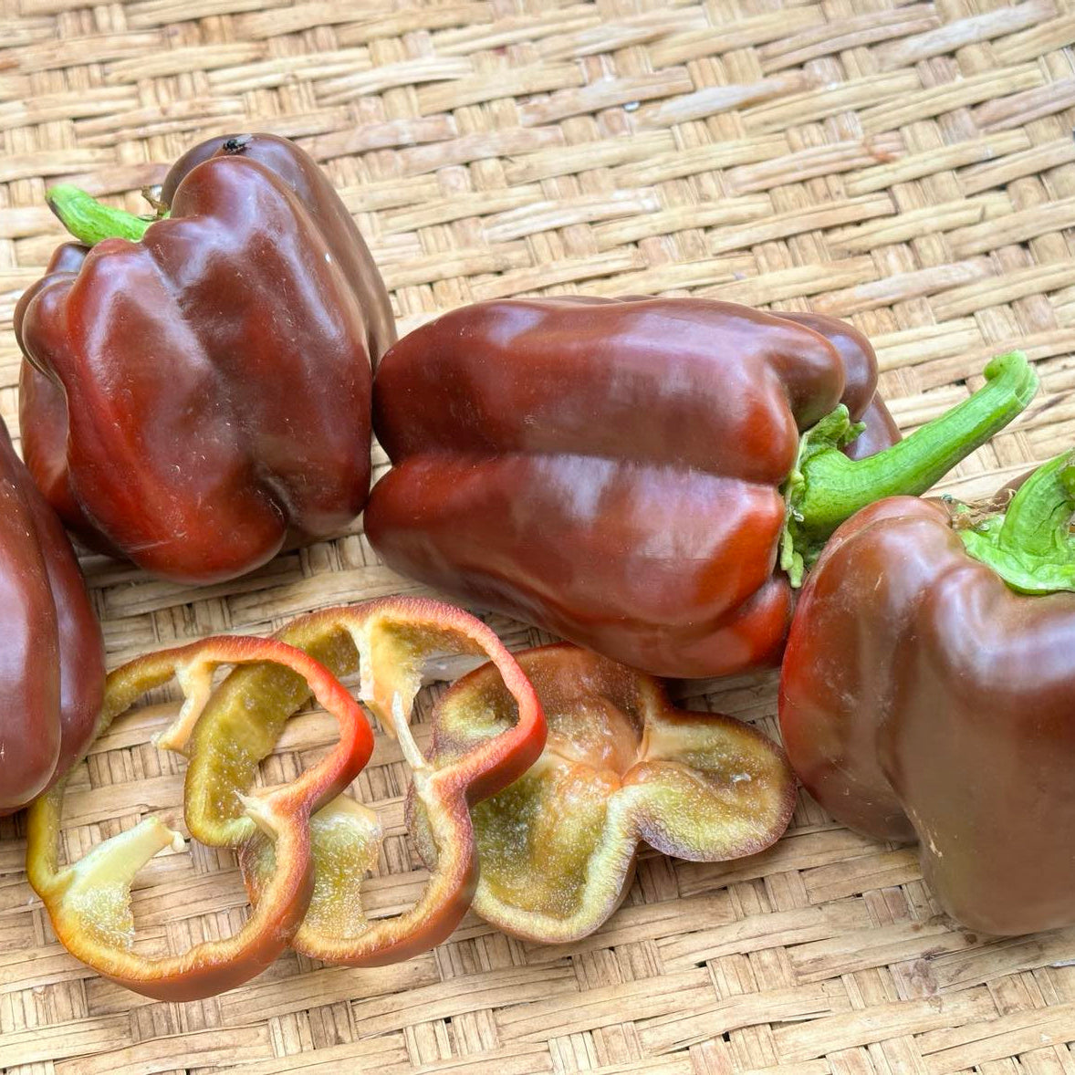 A photograph displaying ripe, deep Chocolate Cake sweet peppers, along with sliced pieces showing the interior, arranged on a woven mat.